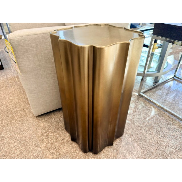 A sleek sculptural accent table in a warm brushed‑gold finish, designed with a softly fluted, petal‑like silhouette that...