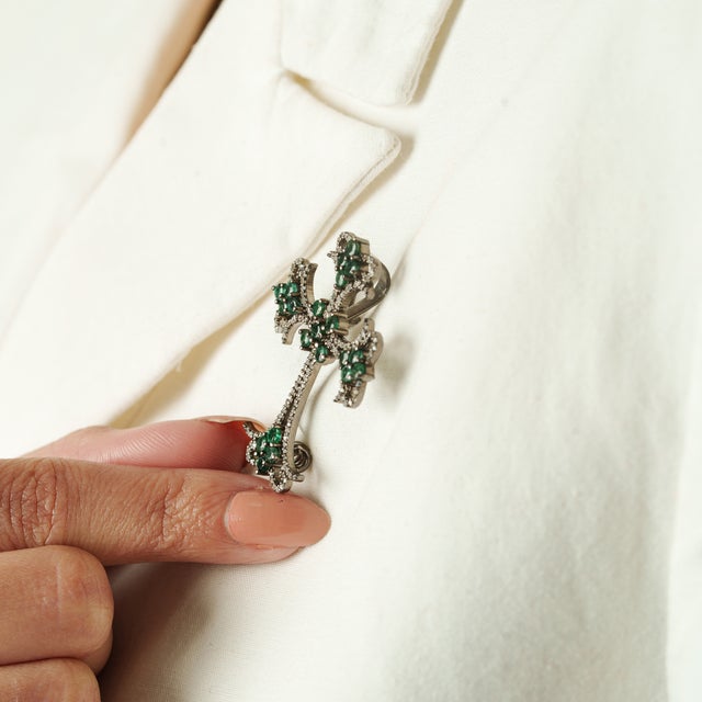 This elegant silver cross brooch features vivid green emeralds accented by sparkling diamonds, arranged in an ornate,...