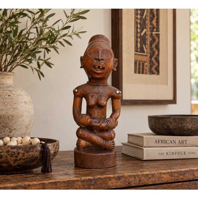 A seated mother holds her infant across her lap, carved in warm reddish-brown wood with a deep, polished glow. She wears a...