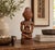 A seated mother holds her infant across her lap, carved in warm reddish-brown wood with a deep, polished glow. She wears a...