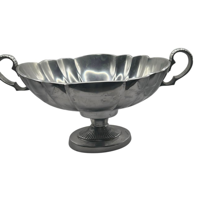 This elegant serving vessels is crafted from pewter and features a fluted body, decorative handles, and a pedestal base....