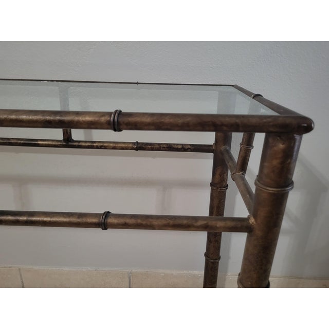 A chic Hollywood Regency–inspired console table featuring a metal frame crafted to resemble bamboo, finished in a warm...