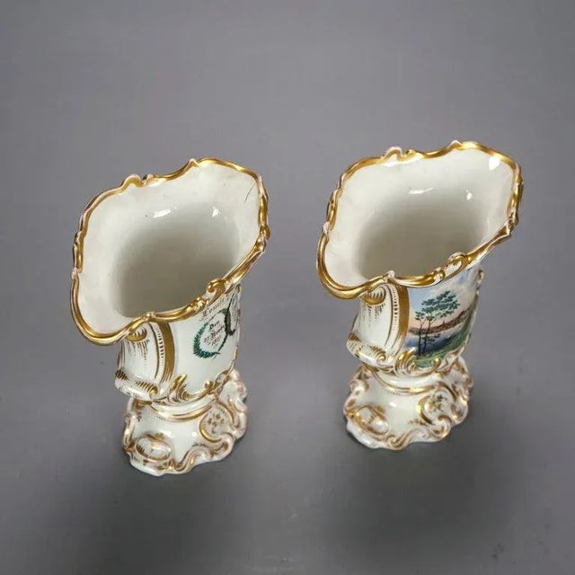 An antique pair of German vases offer porcelain footed construction with scroll form lip over hand painted scenic...