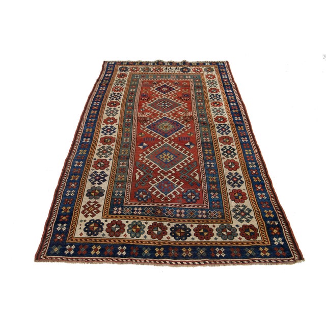 This wool rug features a tribal pattern with beige, blue, and brown accents on a red background inspired by Persian...