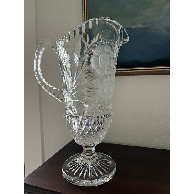 A striking vintage crystal pitcher featuring an elegant pedestal base, dramatic scalloped rim, and intricate cut-glass...