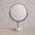 An Italian vanity mirror, circa 1950s. The round glass tilts within a slender brass frame, mounted on a blackened metal...