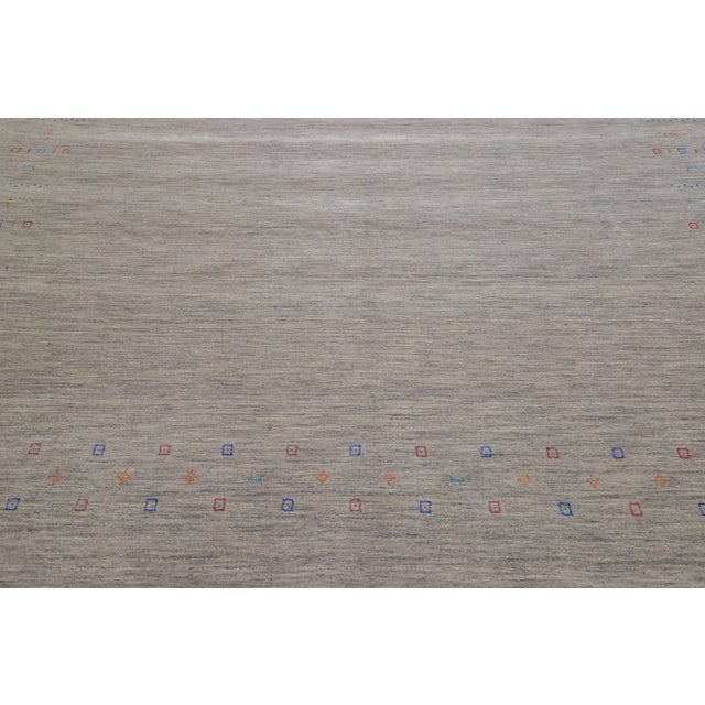 This hand-woven Gabbeh-style wool rug boasts a gray and beige color field featuring a minimalist aesthetic with red, blue,...