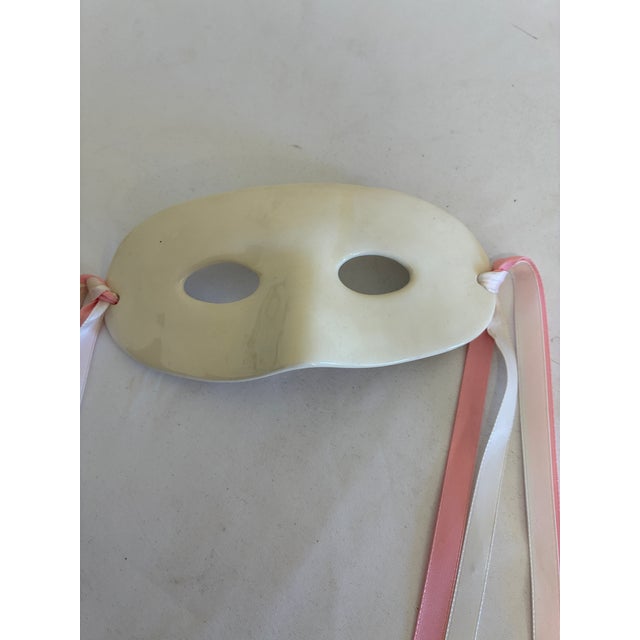 A vintage ceramic masquerade mask featuring a glossy cream finish, oval eye openings, and a softly sculpted nose. Pink and...