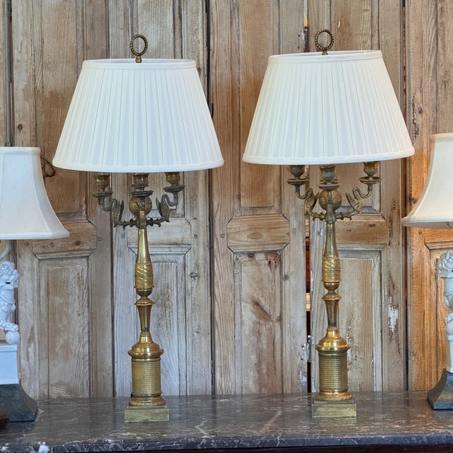 A striking pair of French Empire gilt bronze candelabra, wired as lamps and fitted with pleated white shades. Each has...