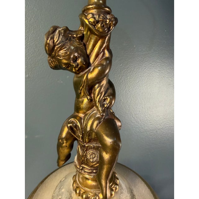 A romantic vintage table lamp featuring a sculptural antique gold-tone cherub / putti figural perched above an ivory-and-...