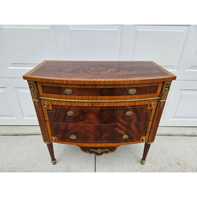 This gorgeous Louis XVI style mahogany chest of drawers features plenty of ormolu or brass-colored appliques. The handmade...