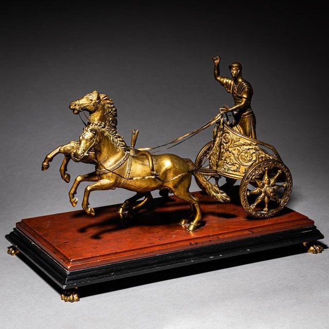 Small chariot in gilt bronze depicting two rearing horses and a charioteer in Roman armor. Mounted on a red and black base...