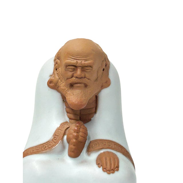 This finely crafted miniature ceramic figurine depicts Damo (Bodhidharma) seated in quiet contemplation, swathed in a...