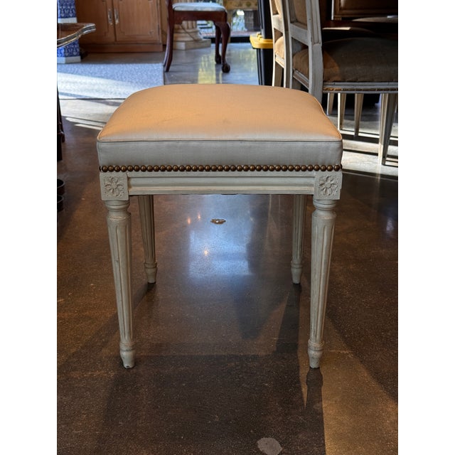Painted stool in the Louis XVI style featuring fluted tapering legs, carved floral corner blocks, and brass nailhead trim...