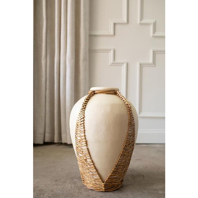 A sculptural vintage terracotta pot encased in an intricately woven rattan frame. The open weave allows the terracotta to...