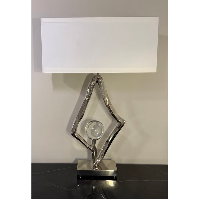 A sculptural Abstract Table Lamp by Global Views featuring an organically contoured polished nickel frame encasing a clear...