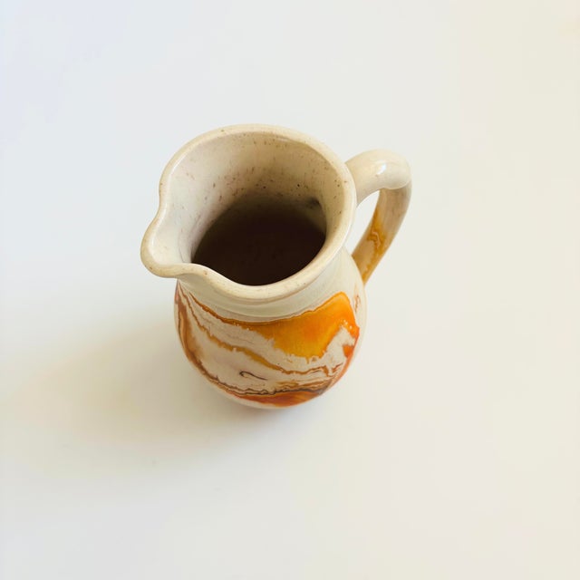 A vintage nemadji pottery pitcher with a beautiful organic pattern of orange swirls on a light off-white background. Matte...
