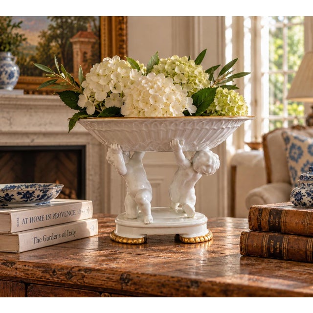 A neoclassical porcelain compote featuring two sculptural putti supporting a fluted bowl, finished with restrained gilt...