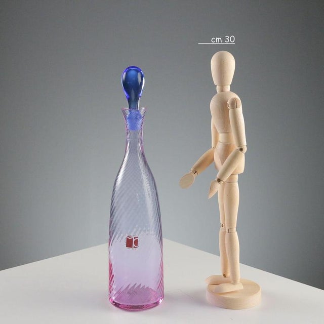Slightly ribbed, spiral-shaped pink glass bottle with a blue cap. Signature of the designer and manufacturer's sticker....