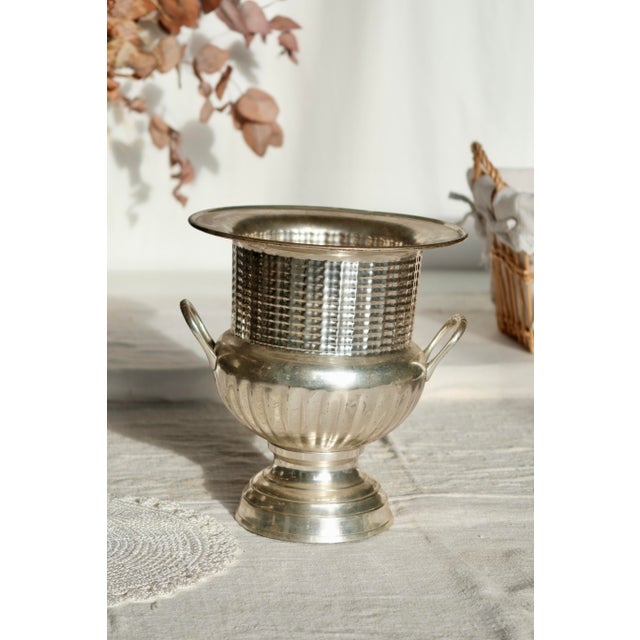 Antique silver-plated champagne bucket, mounted on a wide, elegant base with two delicately designed side handles. The...