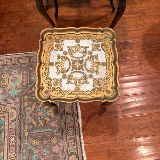 Beautiful set of three French Provincial-style nesting tables featuring intricate gold floral designs, cream-colored tops,...