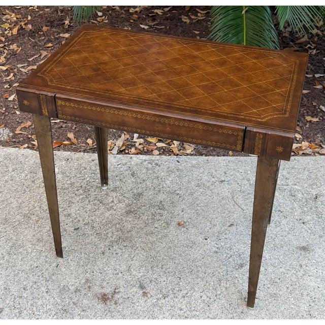 Handsome leather top one drawer table after Maitland Smith. The table top is a faux book. The legs are tapered and metal....