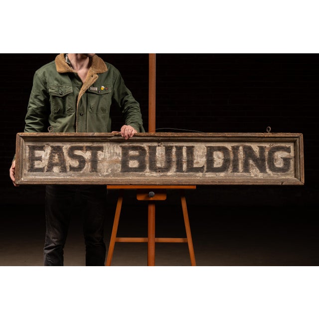 Long, hand-painted wooden directional sign reading "EAST BUILDING," rendered in bold block lettering over a heavily...