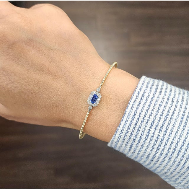 This elegant bangle bracelet embodies a sleek and modern design. At its center, a natural emerald-cut blue sapphire...