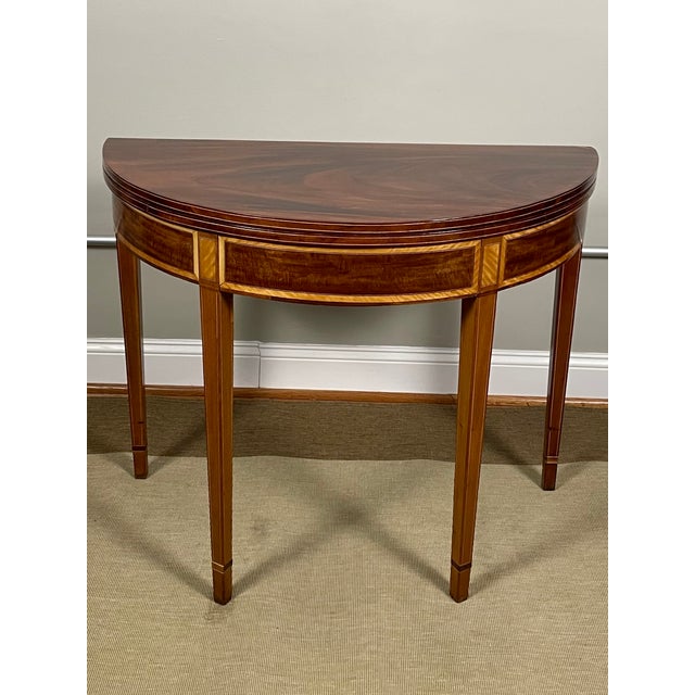 An English George III demilune table featuring a solid figured mahogany top with satinwood pilasters and banding, framed...