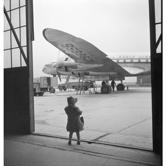 Young Traveller by Toni Frissell A young girl stands in her raincoat awaiting a plane to be refilled for her travels....