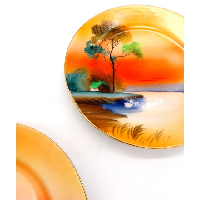 Pair of vintage Noritake porcelain plates, featuring a hand-painted sunset landscape scene. Both plates are finished with...