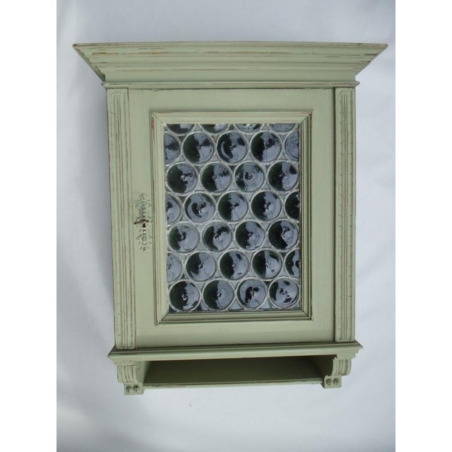 An antique hanging cabinet with a small shelf at the bottom. It's olive green in color and has a shabby chic finish. There...