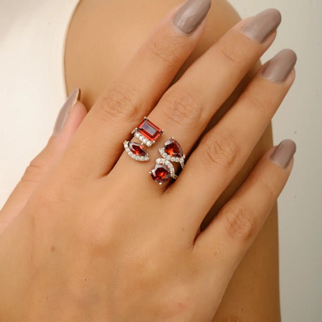 A bold and artistic statement piece featuring four richly hued garnet stones in a mix of striking cuts — emerald, pear,...