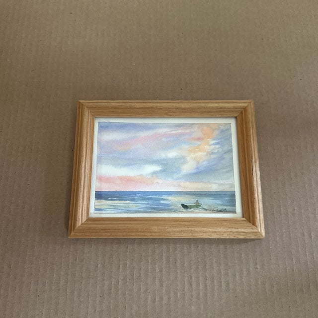 A tranquil seascape in pastel tones of peach and blue, framed in warm wood. A small boat drifts peacefully across quiet...