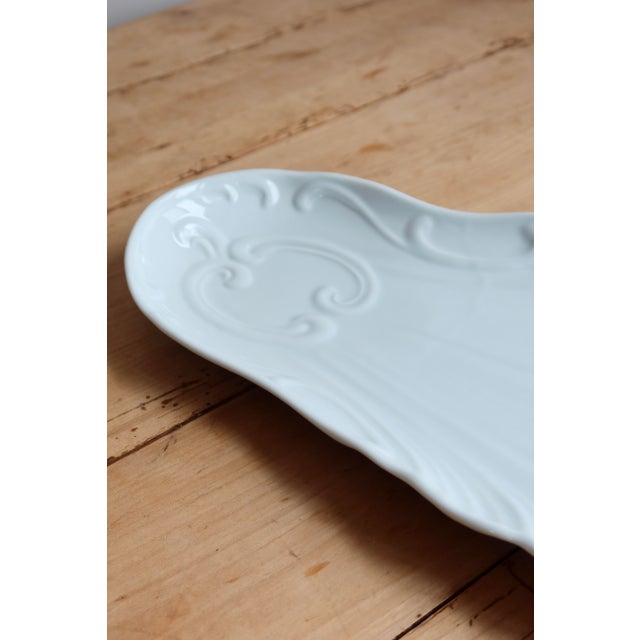 Large white porcelain serving platter in a shell shape with scalloped edges and a raised scroll design. Its elongated...