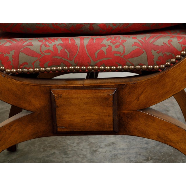 An Austrian Biedermeier period X-form walnut stool from the 19th century, with out-scrolling arms and upholstery. Created...