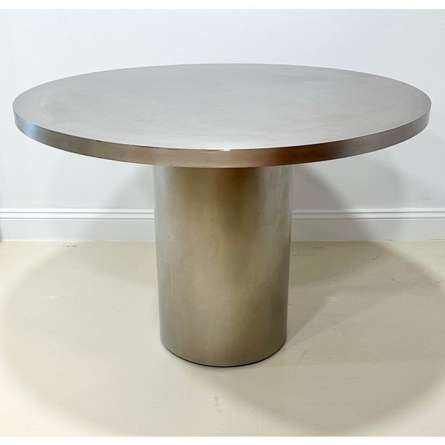 A striking architectural pedestal table crafted in brushed stainless steel, featuring a substantial round top supported by...