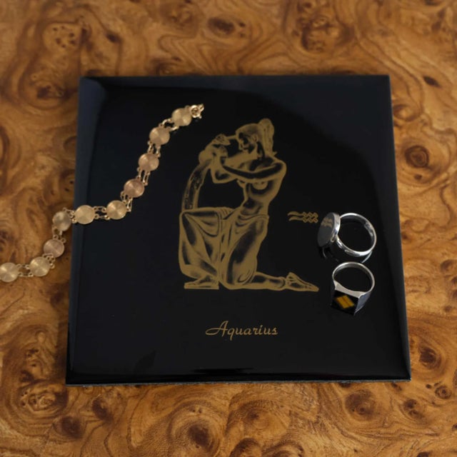 Shiny black decorative ceramic tile features a vibrant gold zodiac print of the Aquarius sign. Tile has a hook on the back...