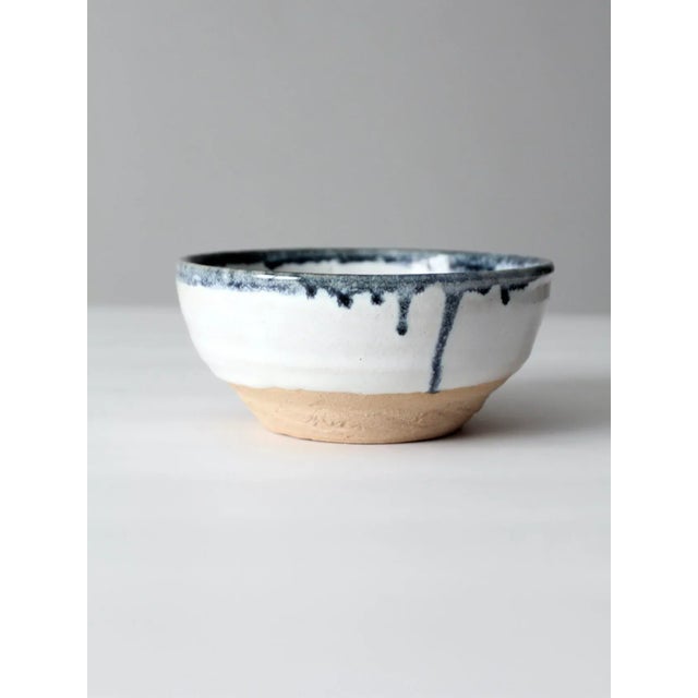 A vintage handmade studio pottery bowl with a soft white glaze and deep blue drip detail at the rim, flowing in natural,...