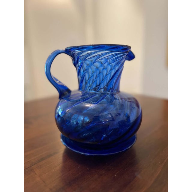 Cobalt blue glass pitcher with a rounded body, twisted ribbed neck, flared rim, and applied handle. Hand-blown, with a...