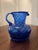 Cobalt blue glass pitcher with a rounded body, twisted ribbed neck, flared rim, and applied handle. Hand-blown, with a...