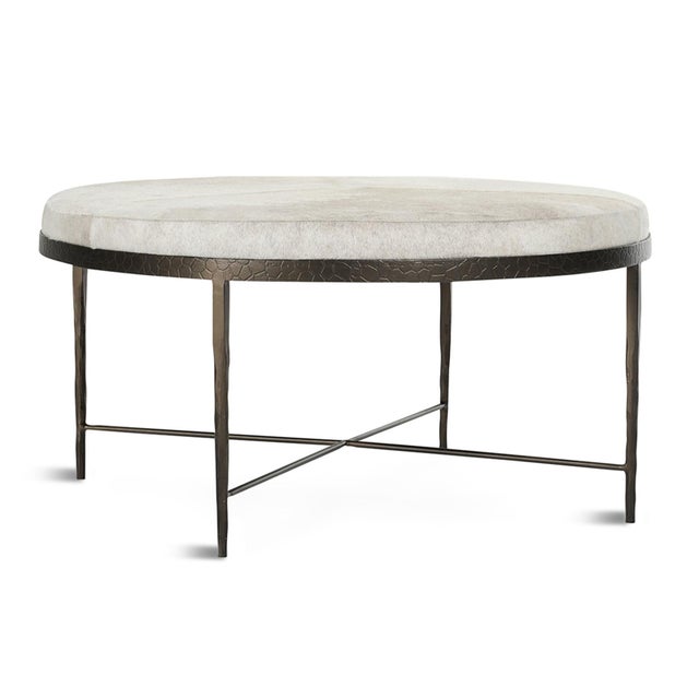 Chic ivory hair-on-hide upholstered round ottoman or coffee table set within an etched iron base. The natural hide...