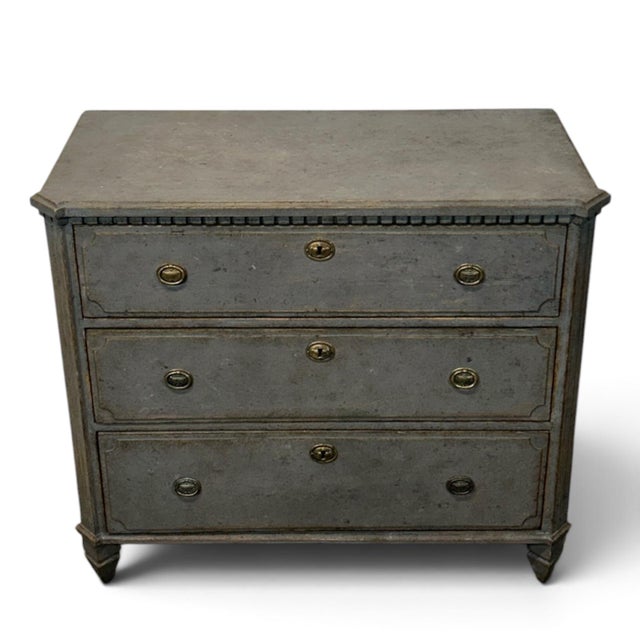A classic Gustavian-style chest of drawers in grey. A decorative toothed trim sits beneath the top. The corners are...