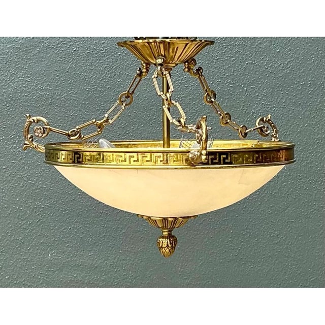 This pendant light presents a wide, shallow bowl of frosted alabaster-toned glass suspended within a substantial gilt-...
