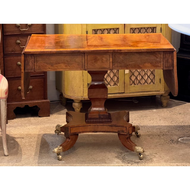 A Regency sofa table in figured wood with crossbanded edges and drop leaves that extend the top. The frieze contains three...