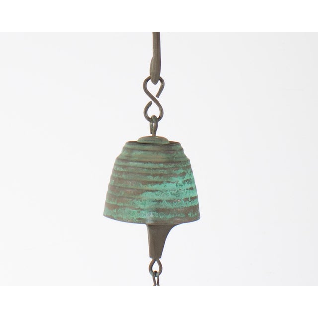 A Harmony hollow bell by American artist Jeff Cross. The metal bell is a greenish-blue and hangs from a curved shape. It...
