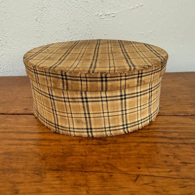Antique rustic round band box with a fitted lid, covered in a tan plaid fabric crossed with black and brown stripes. This...