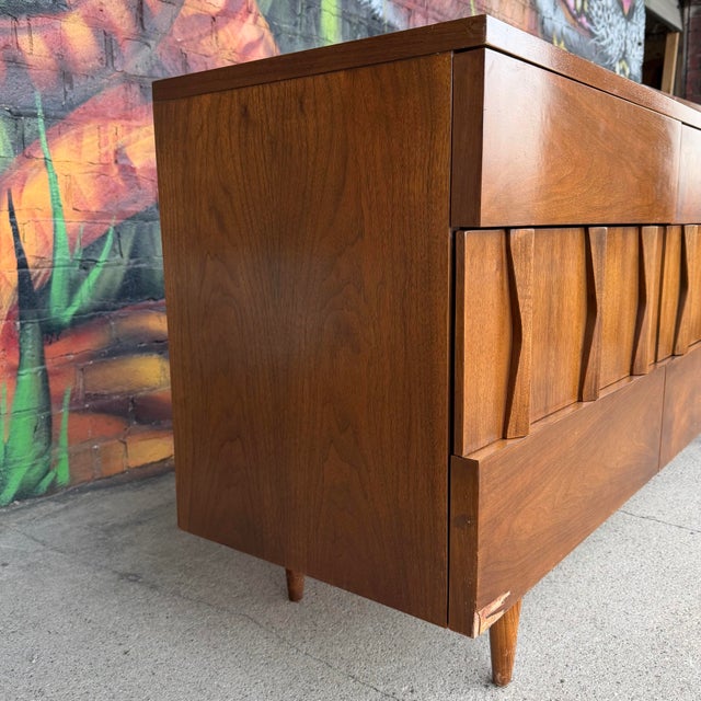This American of Martinsville mid-century modern credenza listing features a walnut wood frame, nine drawers with sculpted...