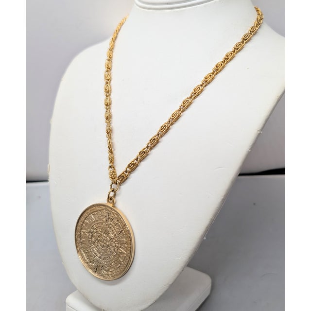 Circa early 1970s goldtone round pendant with Aztec style face in the center and geometric design surround on a textured...