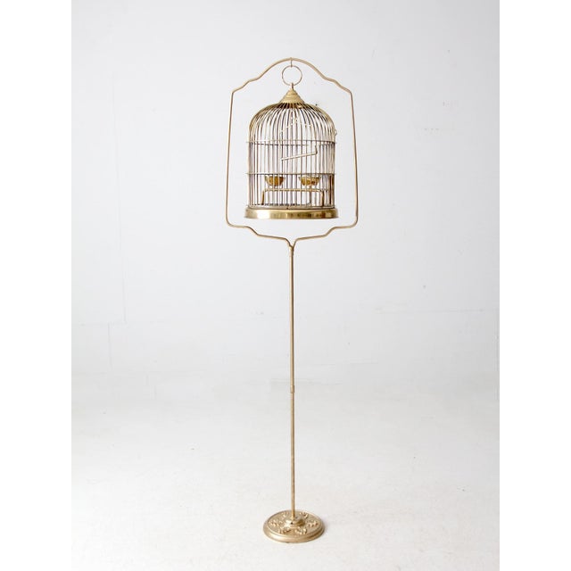 This vintage brass birdcage with stand features a classic pedestal design, two brass feeders, and a hanging perch bar. The...